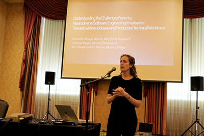 A presenter stands in front of a mic and her powerpoint. A presenter stands in front of a mic and her powerpoint.