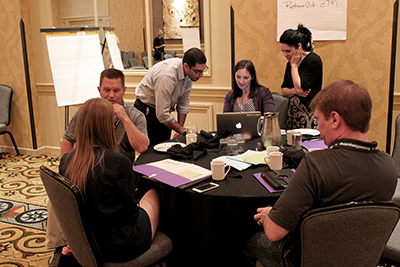 Three participants look at a computer while others speak around the table. Three participants look at a computer while others speak around the table.